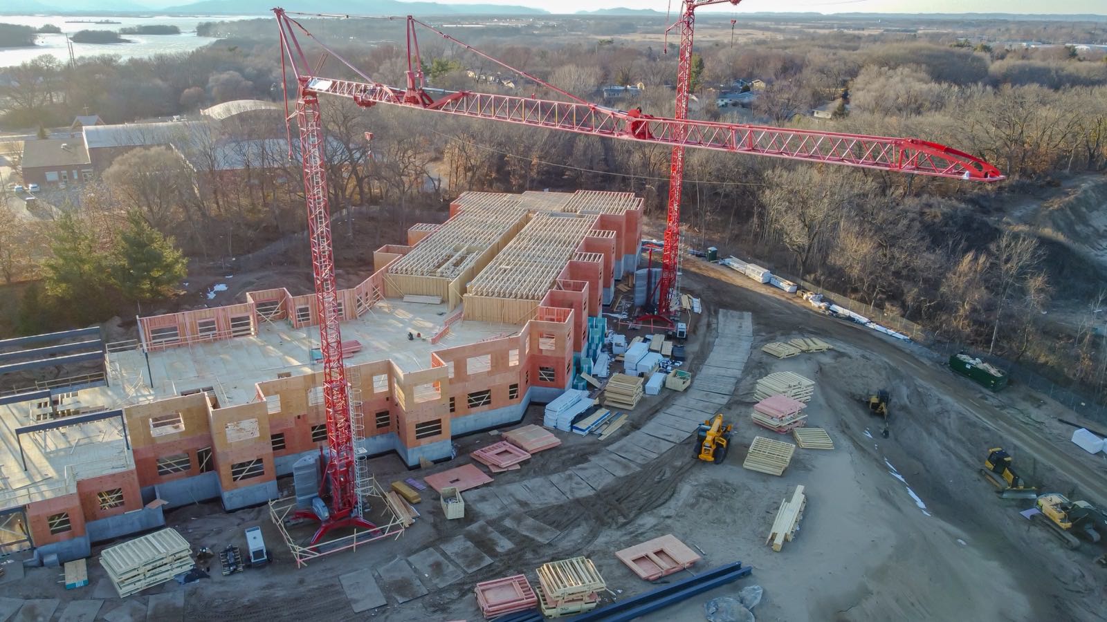 Luftbild einer Baustelle: Zwei rote Turmdrehkrane stehen über einem halbfertigen Gebäude mit Holzrahmenwänden auf Betonsockel, ringsum liegen Stapel vorgefertigter Wandtafeln und stehen gelbe Baumaschinen.