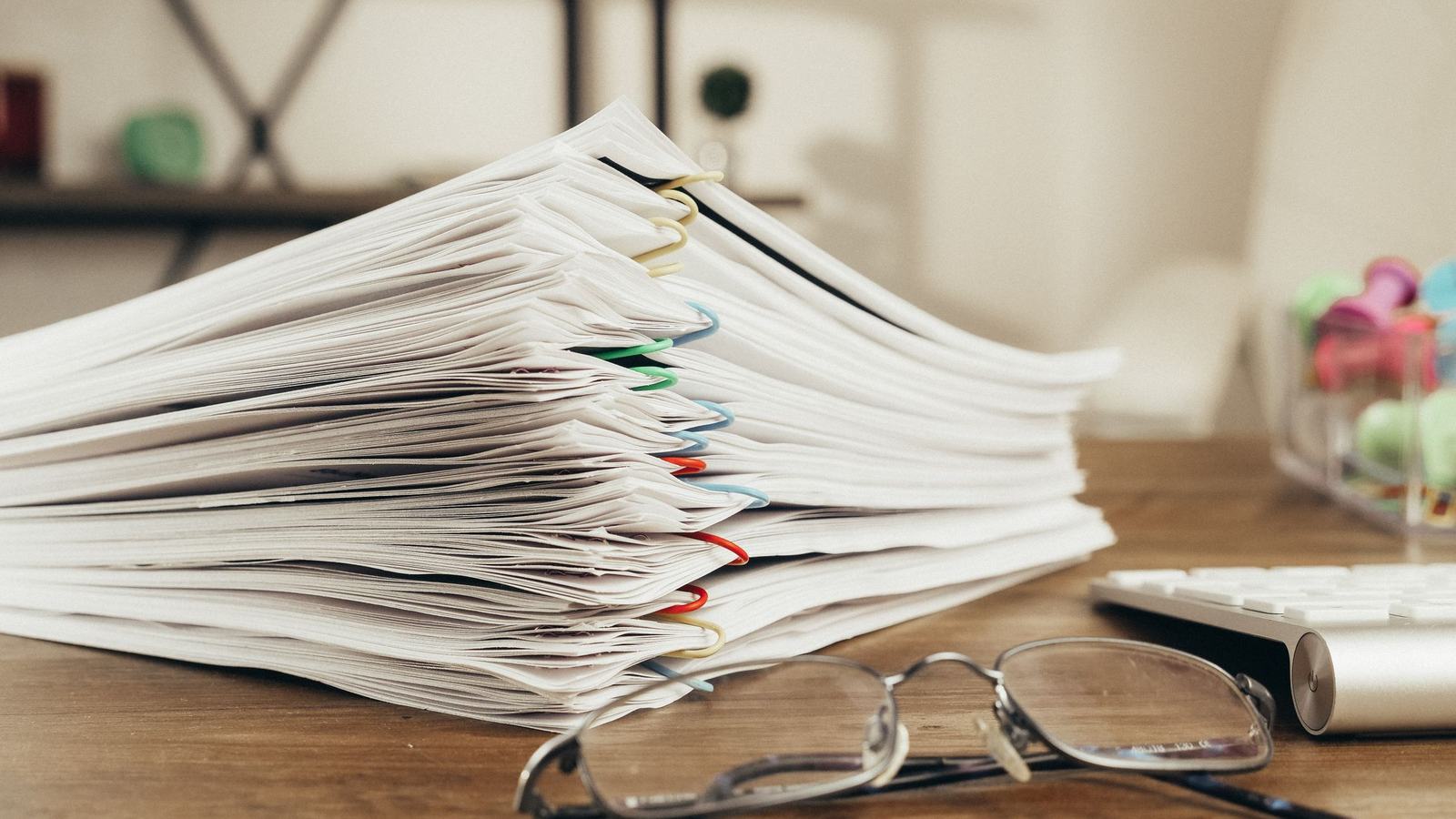 A tall stack of paper documents held together with binder clips, a pair of glasses resting in front of them.