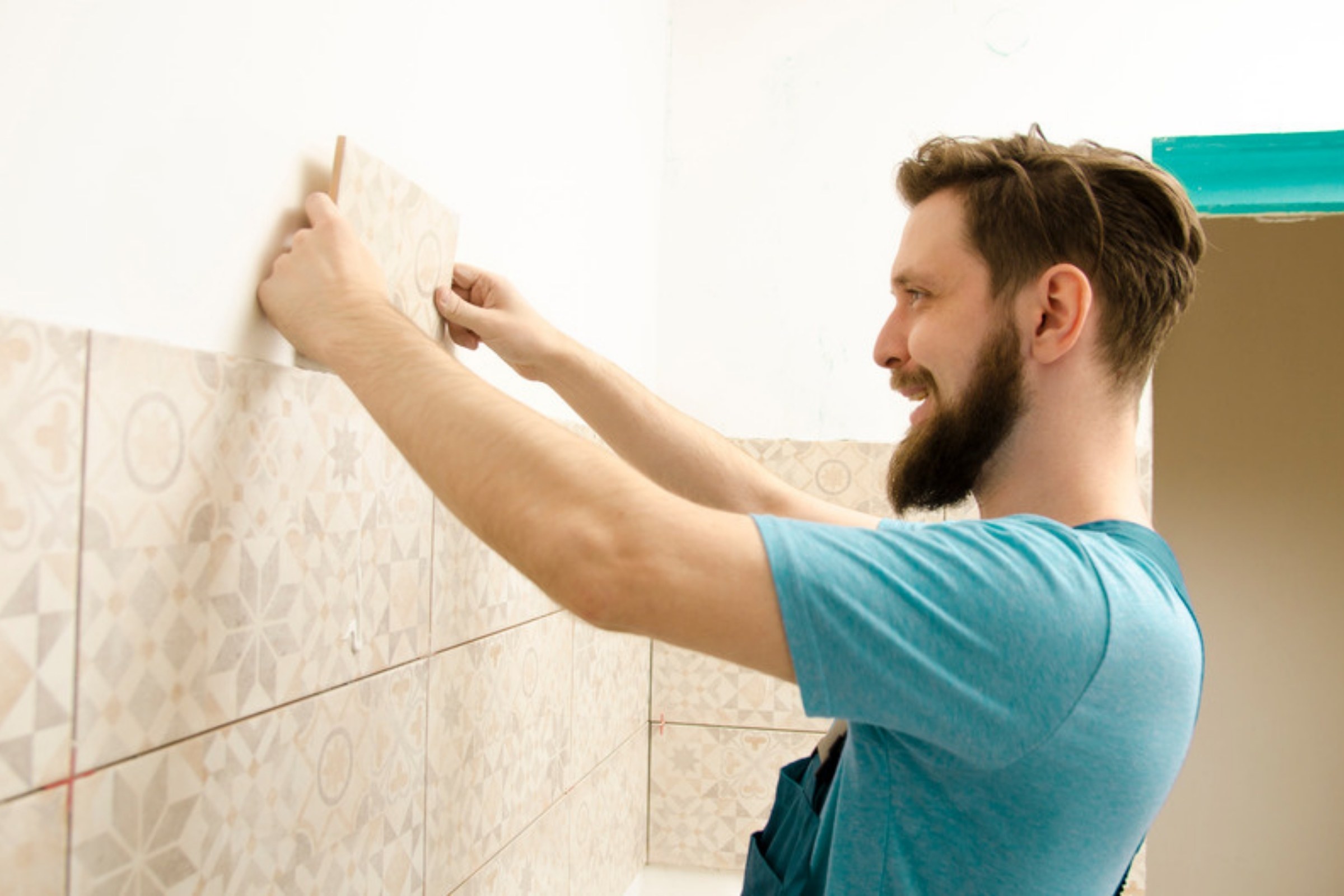 Fliesenleger im blauen Shirt setzt eine gemusterte Wandfliese an, die halbe Wand ist schon gefliest.